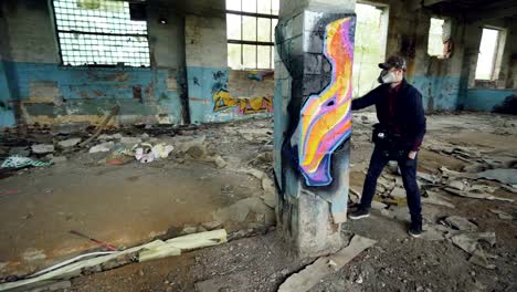 Pan-shot-of-masked-graffiti-artist-drawing-abstract-images-on-pillar-in-large-empty-building-using-paint-spray.-Painter-is-wearing-casual-clothing-and-protective-gloves.