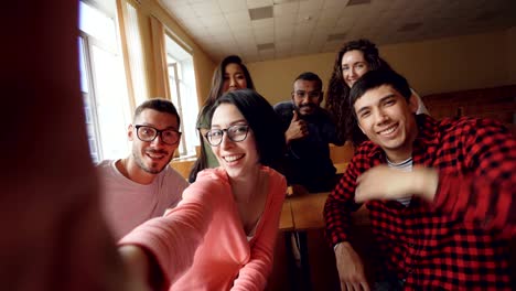 Point-of-view-shot-of-attractive-young-men-and-women-taking-selfie-in-lecture-hall,-posing-with-textbook-and-showing-hand-gestures-and-expressive-faces.