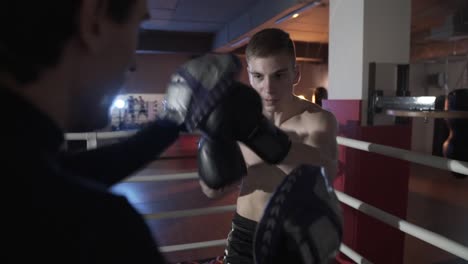 Eine-Nahaufnahme-von-einem-Zeitlupen-Schuss,-ein-Cut-Off,-die-Aufnahme-eines-europäisch-anmutende-Jünglings-mit-Sport-Shorts-mit-einer-nackten-Brust-Boxhandschuhe-anziehen,-die-Technik-des-Kampfes-mit-dem-Coach-arbeitet