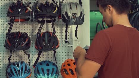 A-young-Caucasian-man-stands-near-a-stand-in-a-bicycle-store-in-the-head-protection-department.-Choosing-a-bicycle-helmet-in-a-small-store