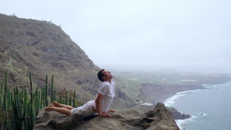 The-man-standing-on-the-edge-of-a-cliff-in-the-pose-of-the-dog-with-views-of-the-ocean,-breathe-in-the-sea-air-during-a-yoga-journey-through-the-Islands