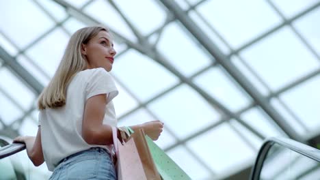 Attractive-young-woman-with-shopping-bags-observing-shopping-mall-and-smiling-while-moving-up-on-escalator-in-slow-motion