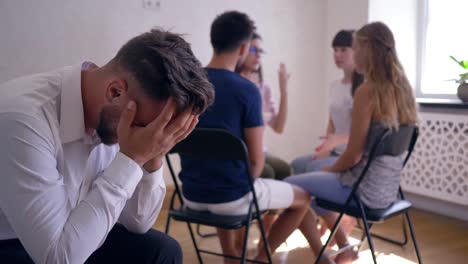 sad-man-crying-and-covers-face-with-hands-on-group-therapy-session-on-background-of-people-sitting-on-chairs-in-circle