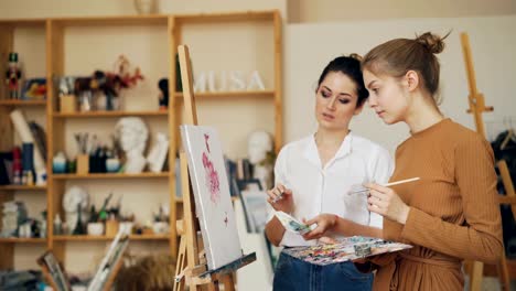 Experienced-painting-teacher-is-talking-to-her-female-student-standing-near-picture-on-easel-and-discussing-artwork.-Shelves-with-tools-are-visible.