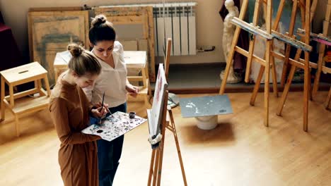 Alto-ángulo-de-visión-de-dos-mujeres-jóvenes-arte-maestra-y-alumno-mezclando-colores-en-la-paleta,-hablando,-sonriendo-y-pintura-sobre-lienzo-en-estudio-de-Artes-modert.