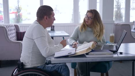 Educación-individual-para-lisiado,-tutor-lección-de-sujeta-gafas-mujeres-para-los-hombres-con-discapacidad-en-silla-de-ruedas-utilizando-un-ordenador-portátil-y-libros