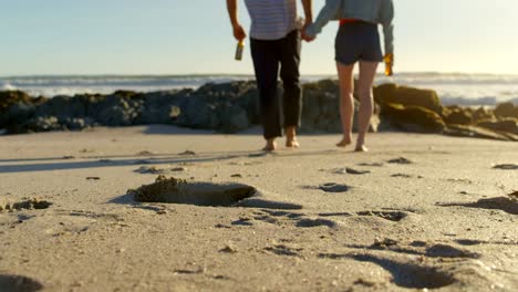 Paar-Hand-in-Hand-mit-Bierflasche-zu-Fuß-am-Strand-4k