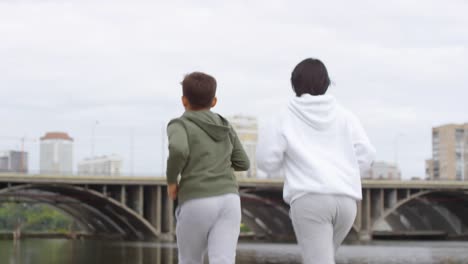 Rear-View-of-Mother-and-Son-Jogging-along-Riverside-in-City