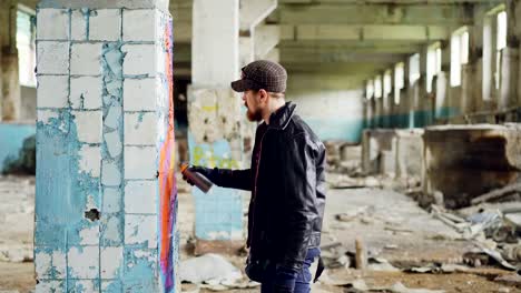 Side-view-of-graffiti-artist-bearded-guy-drawing-on-damaged-column-inside-emply-industrial-building-using-bright-aerosol-paint.-Creativity-and-people-concept.