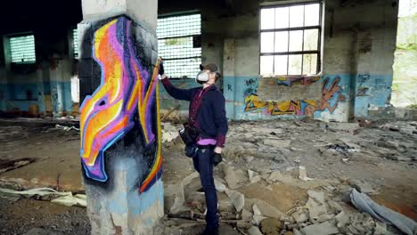 Young-man-professional-graffiti-painter-is-working-inside-abandoned-building,-he-is-painting-with-spray-aerosol-paint-on-high-column.-Modern-art-and-creativity-concept.