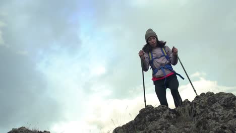 Back-view-of-excited-hipster-girl-standing-on-high-mountain-top-feeling-free-raising-hands-up,-happy-female-traveler-with-backpack-celebrating-achievement-of-reaching-peak-during-hiking-tour