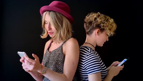 Two-young-beautiful-female-friends-standing-back-to-back-and-typing-on-phone,-isolated-on-background