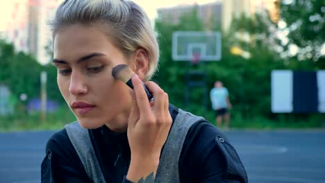 Close-footage-of-beautiful-female-basketball-player-applying-make-up-and-sitting-on-ground,-men-playing-in-background