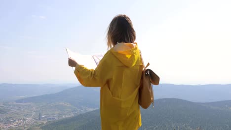 Back-view-of-Caucasian-female-hiker-in-yellow-raincoat-stands-in-the-mountains-with-a-map-in-hand