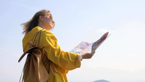 Vista-trasera-del-hiker-mujer-caucásica-en-impermeable-amarillo-se-encuentra-en-las-montañas-con-un-mapa-en-la-mano