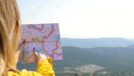 Back-view-of-Caucasian-female-hiker-in-yellow-raincoat-stands-in-the-mountains-with-a-map-in-hand