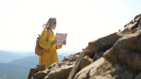 Young-female-hiking-im-yellow-raincoat-with-a-backpack-in-mountains-holding-paper-map-in-hands