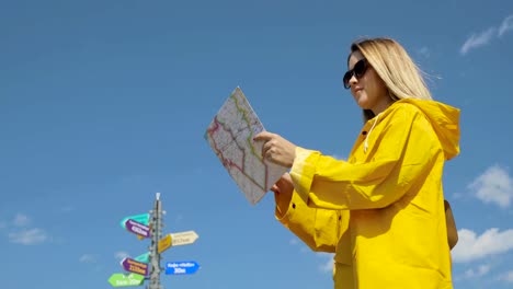 Young-female-hiking-im-yellow-raincoat-with-a-backpack-in-mountains-holding-paper-map-in-hands