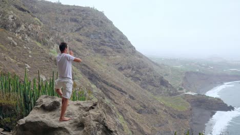 Ein-Mann-steht-am-Rand-einer-Klippe-mit-Blick-auf-den-Ozean-erhebt-seine-Hände-und-atmet-die-Luft-während-yoga