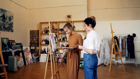 Cheerful-lady-is-painting-picture-together-with-her-female-teacher-professional-artist-using-brush-and-oil-paints.-Modern-workshop-with-easels-and-artworks-is-visible.