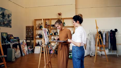 Fröhliche-Dame-ist-Malerei-Bild-zusammen-mit-ihrer-Lehrerin-professioneller-Künstler-mit-Pinsel-und-Ölfarben.-Moderne-Werkstatt-mit-Staffeleien-und-Kunstwerken-ist-sichtbar.