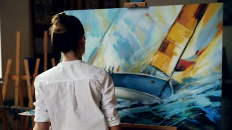 Serious-female-artist-is-working-at-picture-using-oil-paints-and-palette-knife-creating-beautiful-marine-landscape-on-canvas.-Painting-technique-and-painters-concept.