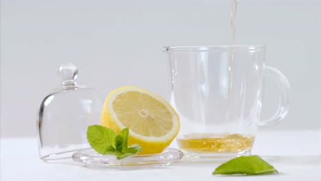 Herbal-tea-being-poured-into-transparent-teapot,-lemon-and-mint-leaves-around,-75-fps-slow-motion