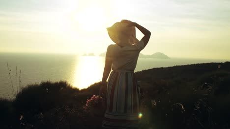 Beautiful-young-woman-wearing-fashion-colorful-dress-with-skirt-and-hat-standing-with-flowers-in-basket-at-sunset-on-Ponza-Island-mountain-coast-Italy.