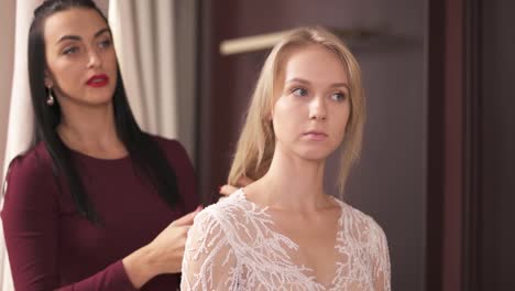 A-young-blonde-in-an-embroidered-fancy-dress-and-a-designer-standing-behind-her-in-the-atelier.-The-designer-stretches-her-hair,-adjusts-the-fabric-on-shoulder-seams.-They-are-talking-and-smiling.