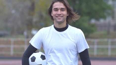 Portrait-of-a-handsome-guy-in-T-shirt-smiling-and-looking-at-the-camera-while-holding-the-ball-in-his-hands-A-young-guy-with-a-soccer-ball-smiles-beautifully-at-the-camera-and-shows-thumb-up-Sporty-young-man-happy-outdoors-Healthy-lifestyle