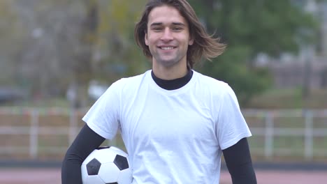 Retrato-de-un-chico-guapo-en-camiseta-sonriendo-y-mirando-a-la-cámara-mientras-sostiene-la-bola-en-su-joven-manos-A-con-un-balón-de-fútbol-sonríe-maravillosamente-en-la-cámara-y-muestra-pulgar-feliz-estilo-de-vida-saludable-al-aire-libre-deportivo-jov