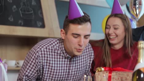 Handsome-man-blowing-birthday-candles-while-close-friends-throwing-a-surprise-party,-slowmotion-in-cozy-modern-apartment