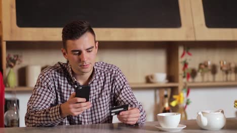 Handsome-man-making-purchase-online,-typing-credit-card-details-on-black-smartphone-in-the-modern-kitchen