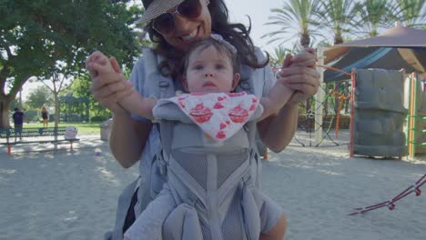 Woman-smiling-at-camera-as-she-holds-baby-in-carrier-at-playground