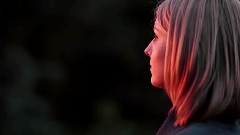 Attractive-young-woman-standing-at-park-in-the-rays-of-the-setting-sun-and-looking-into-the-distance.-Close-up-Side-view