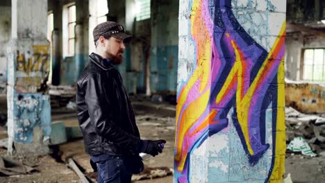 Professional-graffiti-painter-is-creating-abstract-image-on-large-pillar-inside-abandoned-building-with-spray-paint.-Young-man-is-wearing-leather-jacket,-cap-and-gloves.