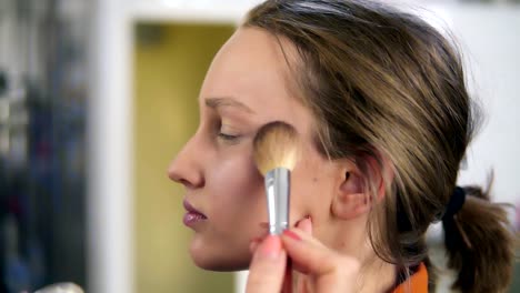 Side-view-of-a-young,-pretty-female-model.-Make-up-process.-Artist-putting-some-transparent,-shiny-rouge-on-woman's-cheek-using-a-brush