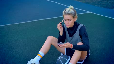 Schöne-junge-blonde-weibliche-Basketballspieler-Make-up-zu-tun-und-sitzen-auf-dem-Boden-mit-Ball,-Sportkleidung-tragen