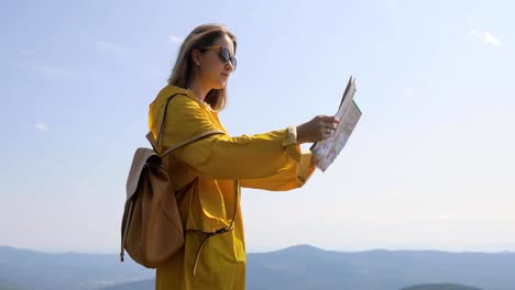 Mujer-joven-en-un-sendero-de-montaña,-de-senderismo-en-impermeable-se-detiene-y-comprueba-el-mapa-para-direcciones.-Excursionista-alcanza-la-cima-de-la-montaña,-comprueba-las-direcciones-en-el-mapa