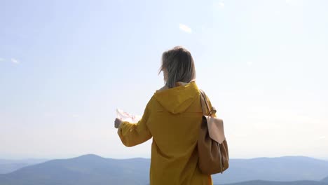 Mujer-joven-en-un-sendero-de-montaña,-de-senderismo-en-impermeable-se-detiene-y-comprueba-el-mapa-para-direcciones.-Excursionista-alcanza-la-cima-de-la-montaña,-comprueba-las-direcciones-en-el-mapa