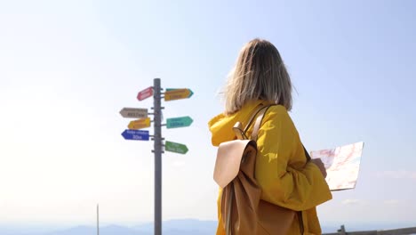 Back-view-of-Caucasian-female-hiker-in-yellow-raincoat-stands-in-the-mountains-with-a-map-in-hand