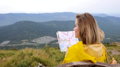 Young-female-hiking-im-yellow-raincoat-with-a-backpack-in-mountains-holding-paper-map-in-hands