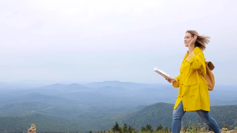 Young-female-hiking-im-yellow-raincoat-with-a-backpack-in-mountains-holding-paper-map-in-hands