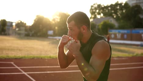 Bearded,-concentrated-man-boxer-doing-boxing-exercise,-warming-up-while-standing-on-stadium-outdoors-in-summer-sun-rays.-Side-view