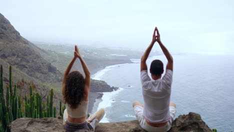 Un-hombre-y-una-mujer-sentada-en-la-cima-de-una-montaña-mirando-el-mar-sentado-en-una-piedra-de-meditación-levantando-sus-manos-y-realizando-una-respiración-relajante.-Islas-Canarias