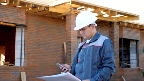 Man-in-white-hardhat-and-overall-holding-paper-plans-of-building-and-using-phone-outside-on-site