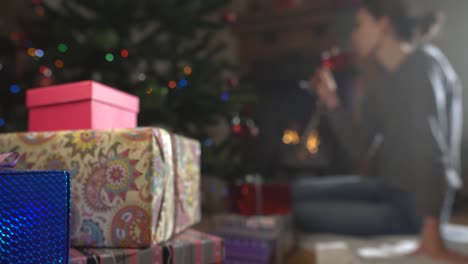 joven-mujer-está-bebiendo-vino-cerca-del-árbol-de-Navidad-decorado-y-junto-a-la-chimenea.-centrarse-en-los-regalos