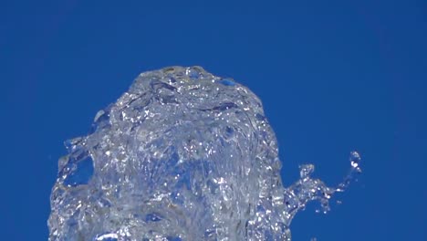 A-splash-of-clear-ice-water.-Closeup-of-light-splashing-water-in-sunny-fountain-in-park-in-summer-on-blue-sky-background.--Water-flow-of-fountain-fly-up-in-air-with-many-splashes.-Slow-motion.