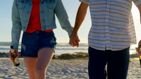 Mid-section-of-couple-holding-hands-with-beer-bottle-walking-on-beach-4k