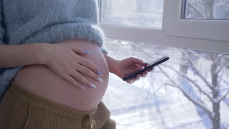 expectant-mom-stroking-her-naked-tummy-and-uses-cellphone-while-standing-by-a-large-window-at-apartament